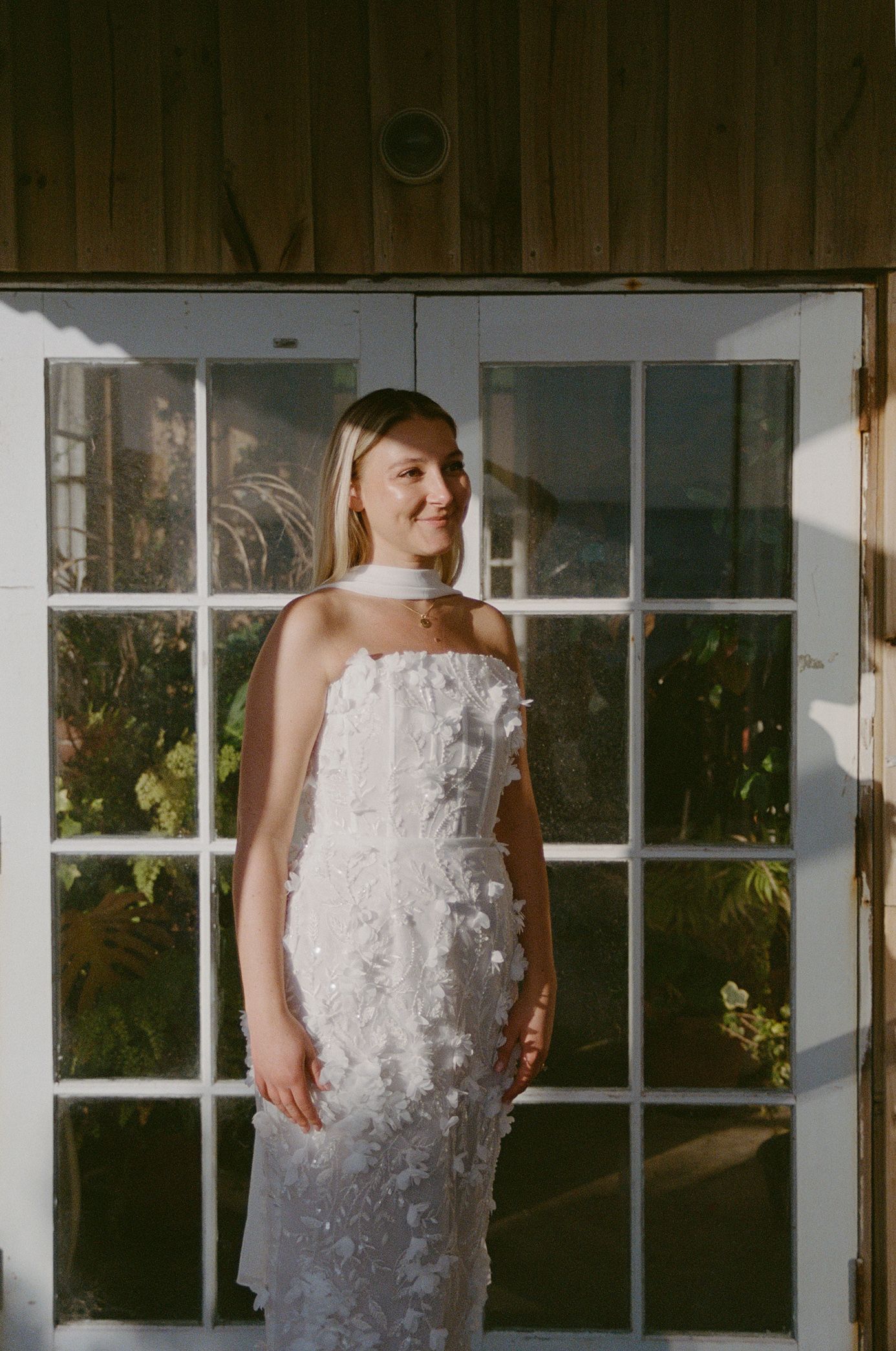 Bride in strapless white floral appliqué wedding gown standing before French doors with sunlight streaming through.