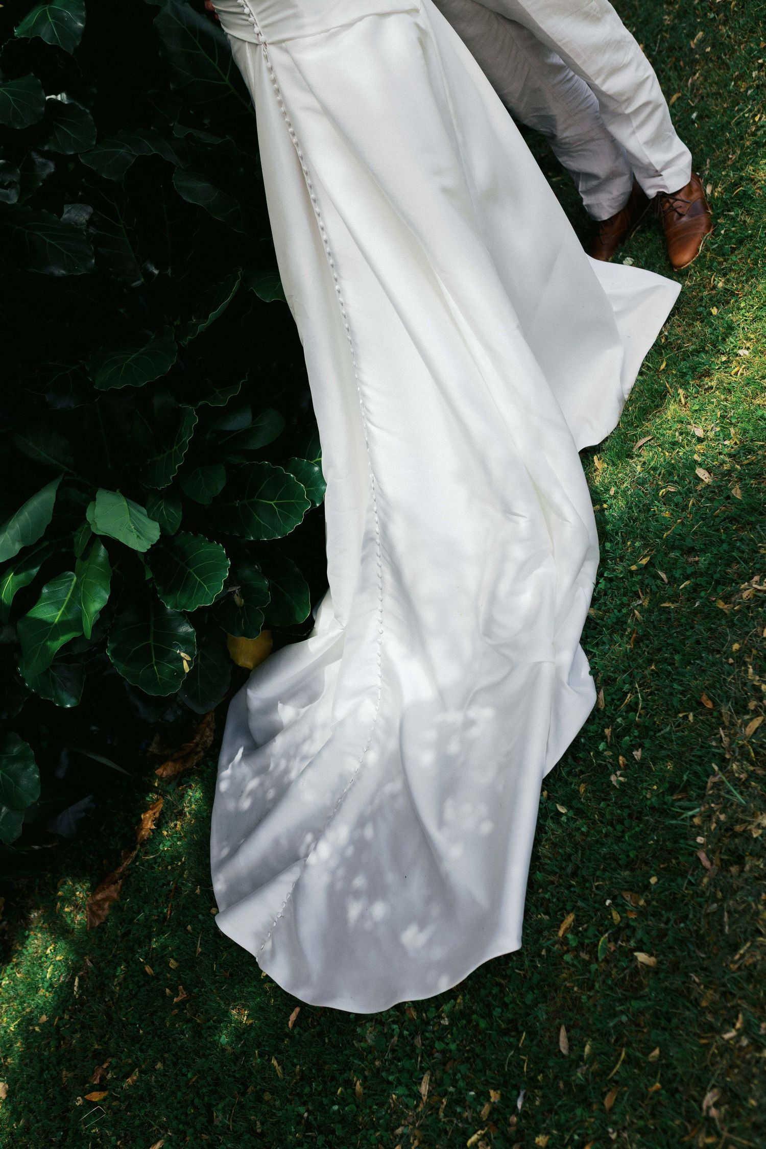 A flowing white wedding dress train trails on green grass beside garden foliage.