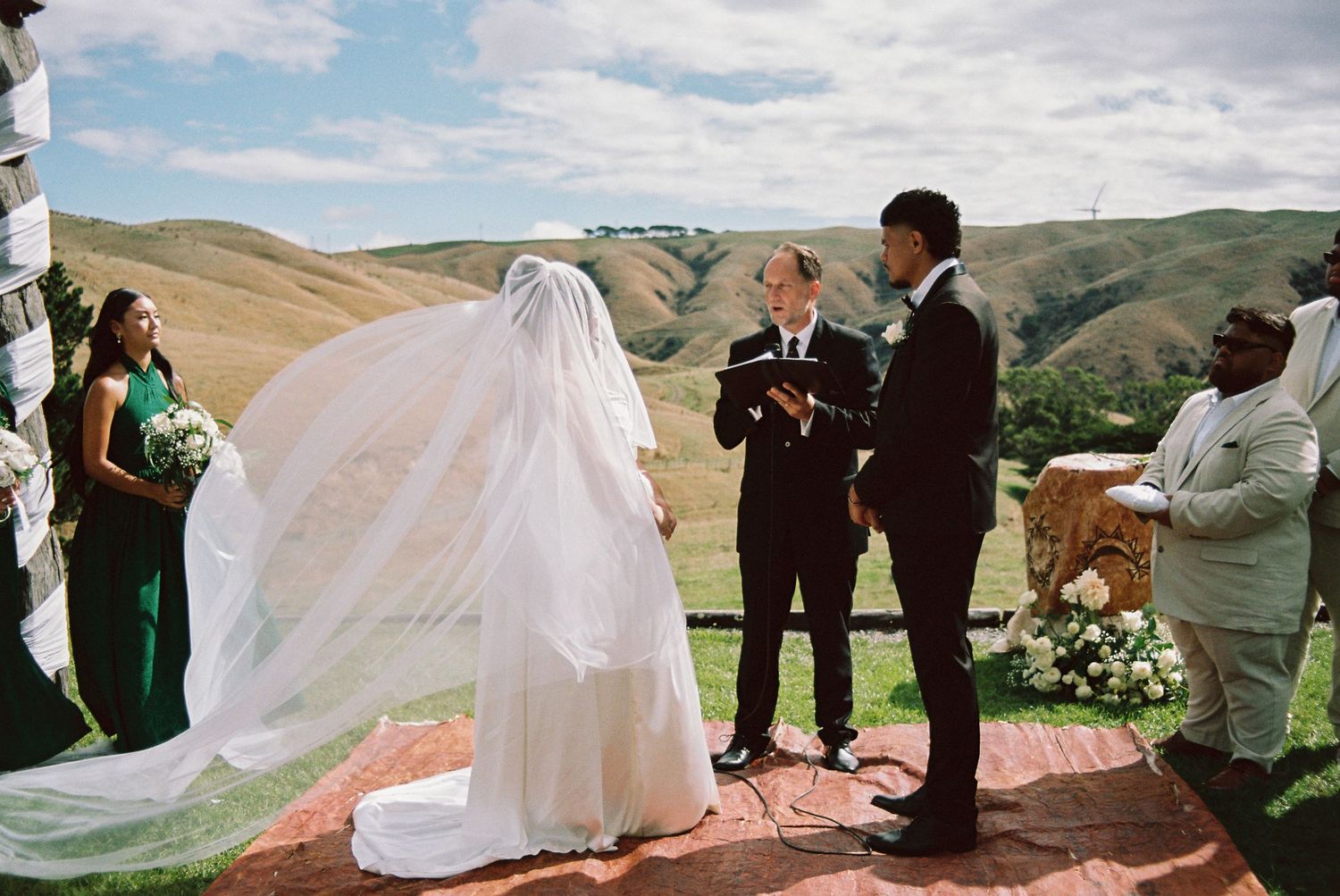 A flowing wedding veil catches the wind dramatically against a scenic mountain landscape backdrop during an outdoor ceremony.