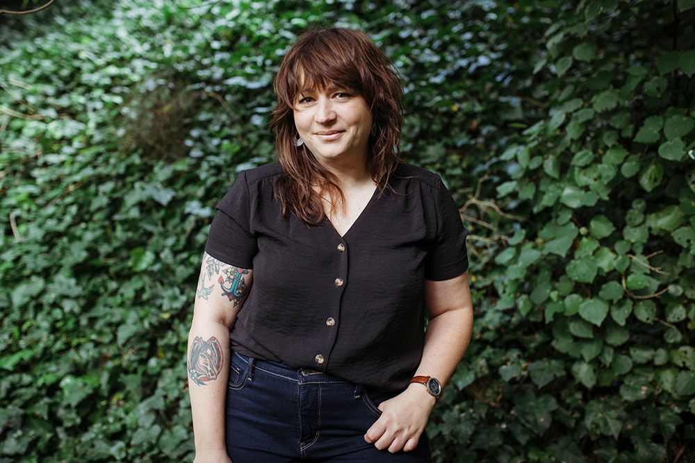 A person in a black shirt smiles warmly while sitting outdoors surrounded by lush green foliage in a natural setting.