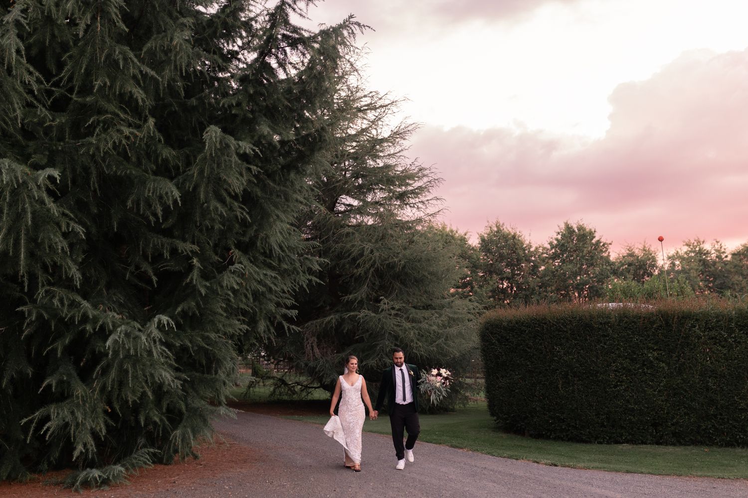 A couple walks hand in hand down a garden path at sunset with evergreen trees in background.