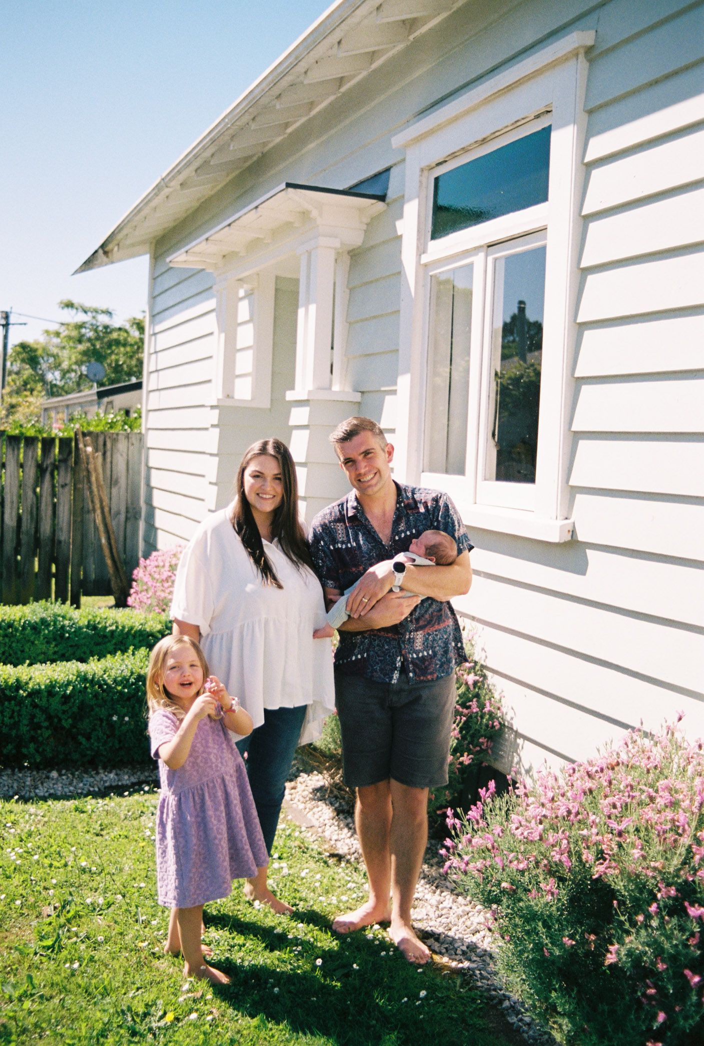 A series of sunny outdoor shots showing loving family interactions beside a white house with pink flowering bushes.