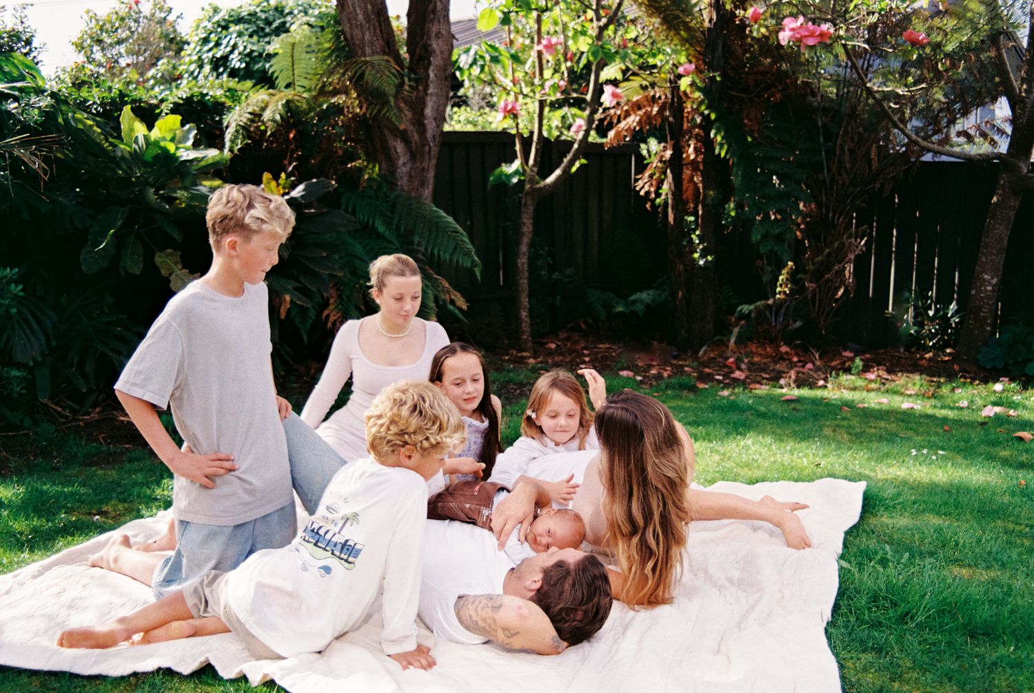 A family lounges together on a blanket in a sunny backyard garden setting during a casual outdoor photoshoot.