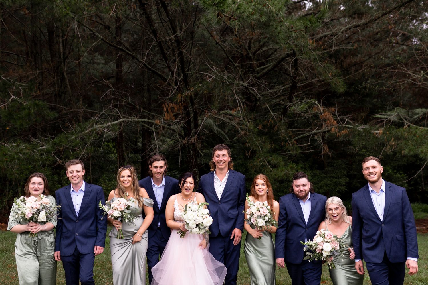 Wedding party in navy and sage poses together for formal group photo in front of pine trees.