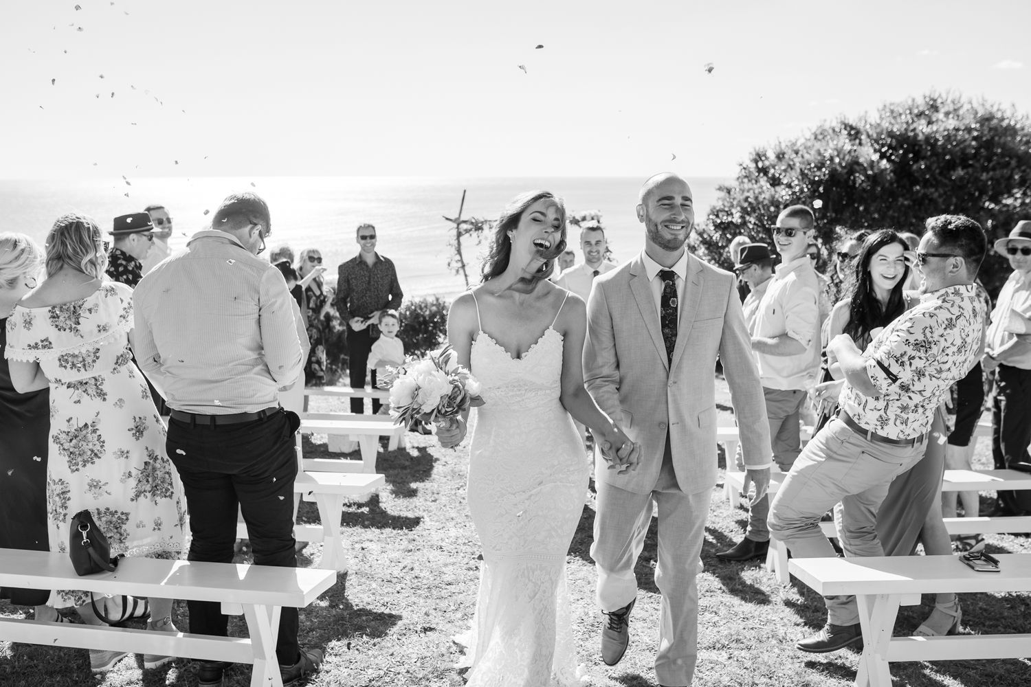 Black and white photo of wedding guests celebrating at an outdoor ceremony with wooden benches.
