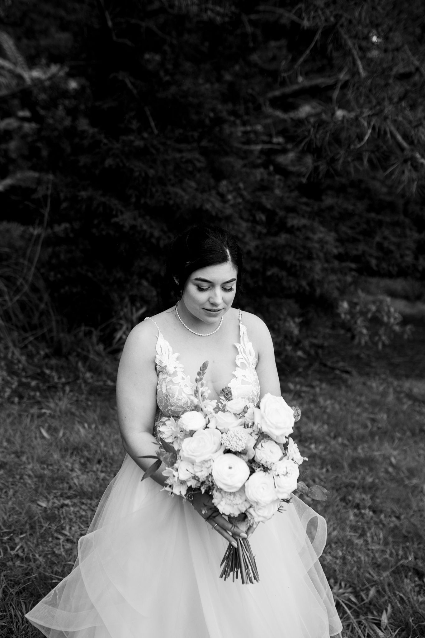 Black and white portrait of bride holding elegant white floral bouquet against dark forest background.