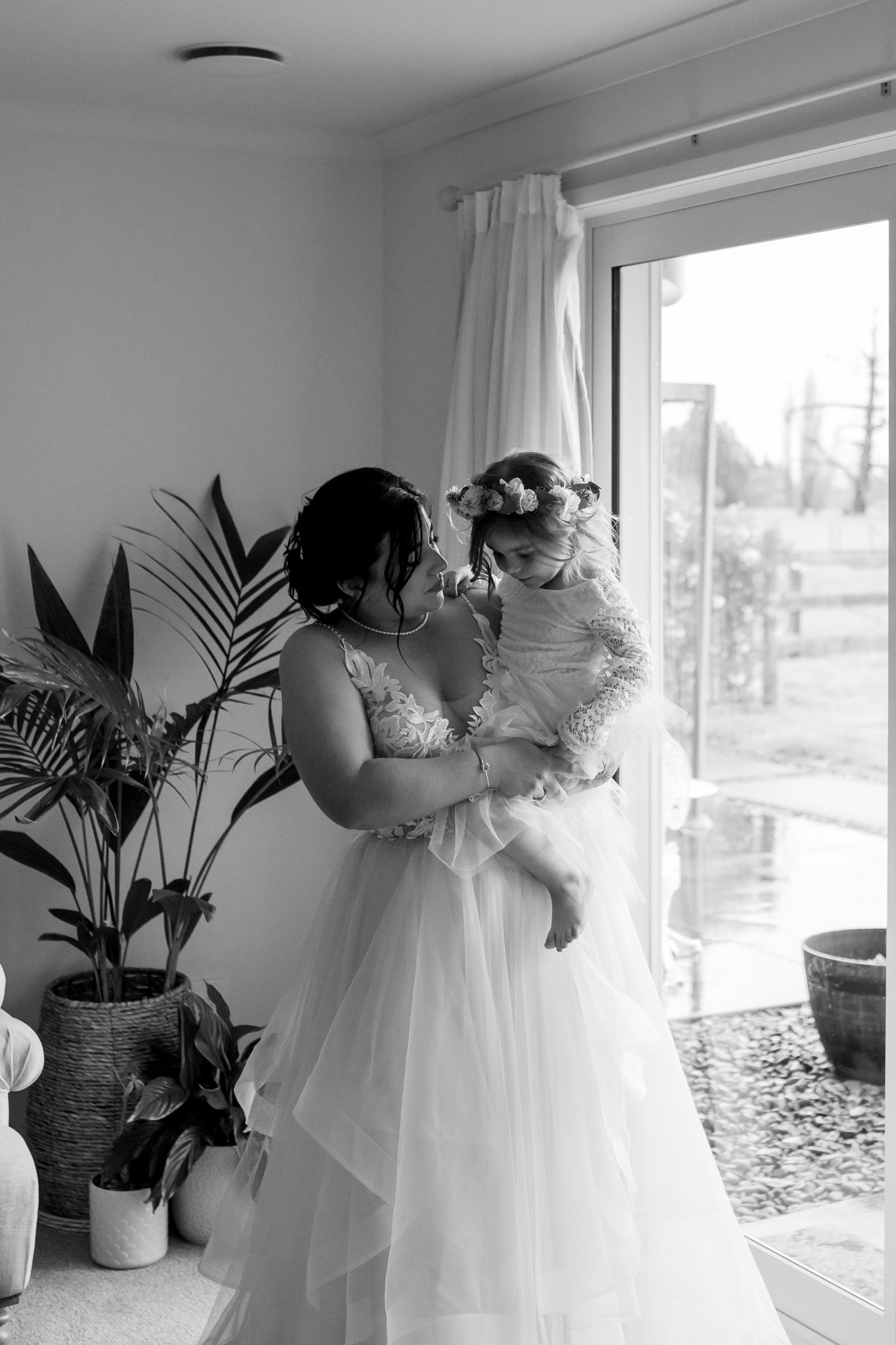 A tender moment in a white wedding dress beside a window with a potted palm plant.