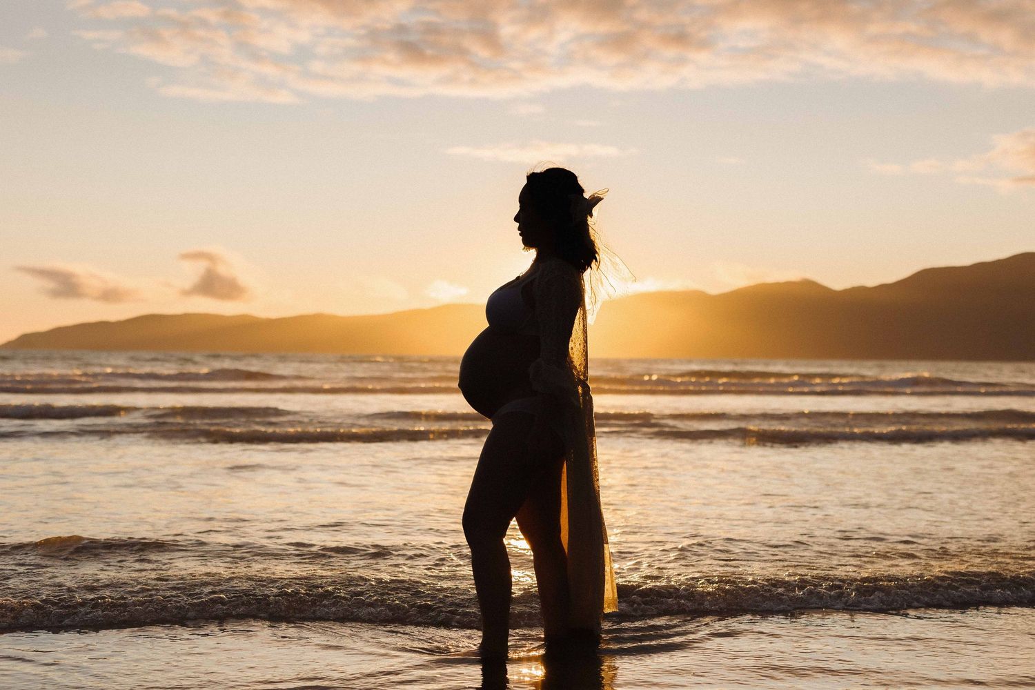 Silhouette of pregnant woman standing on beach at sunset with mountains in background and waves rolling in.