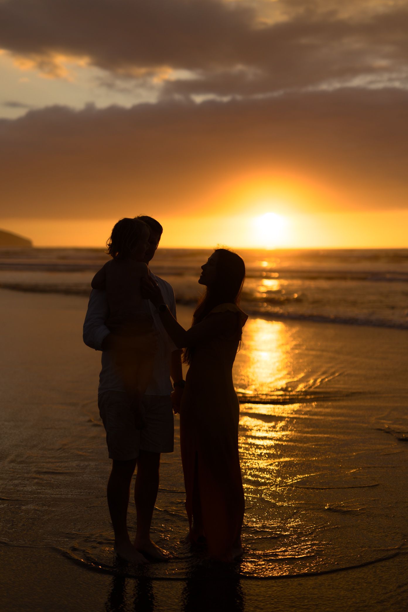 Two silhouetted figures stand together on a beach at sunset as golden light reflects off the ocean waves.