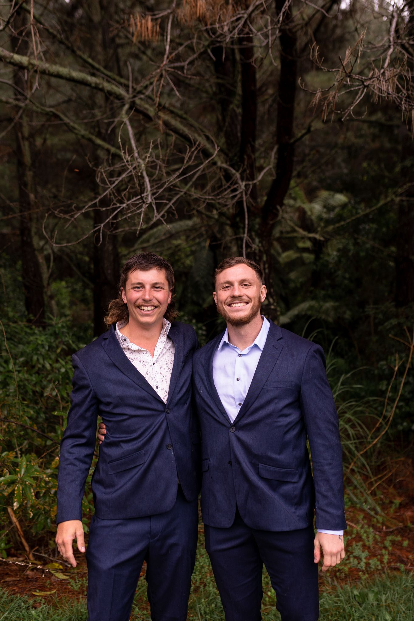 Two people in matching navy suits share a happy moment together in a forest setting.