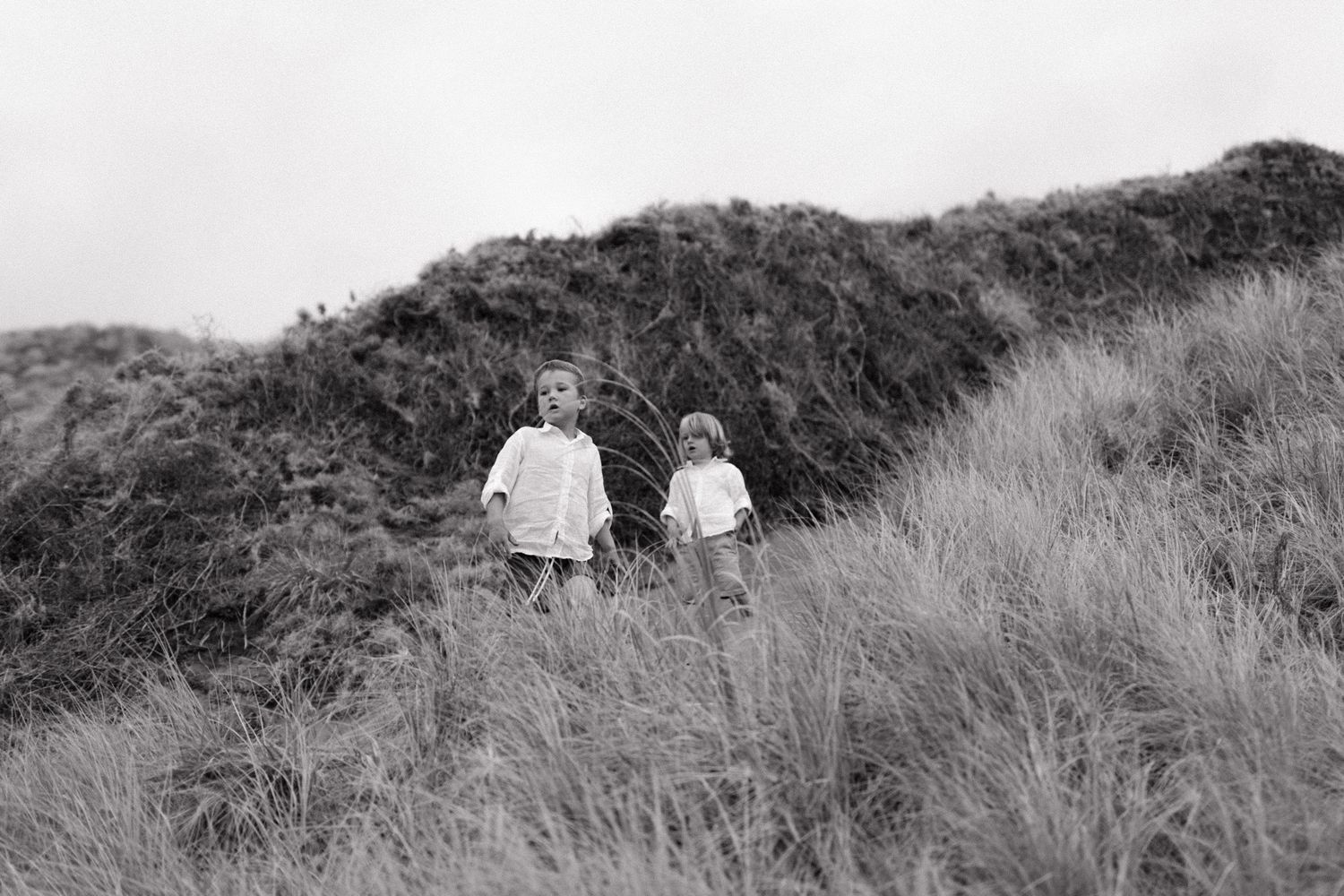 Two people walk together along a grassy hillside path overlooking the ocean on a cloudy day.