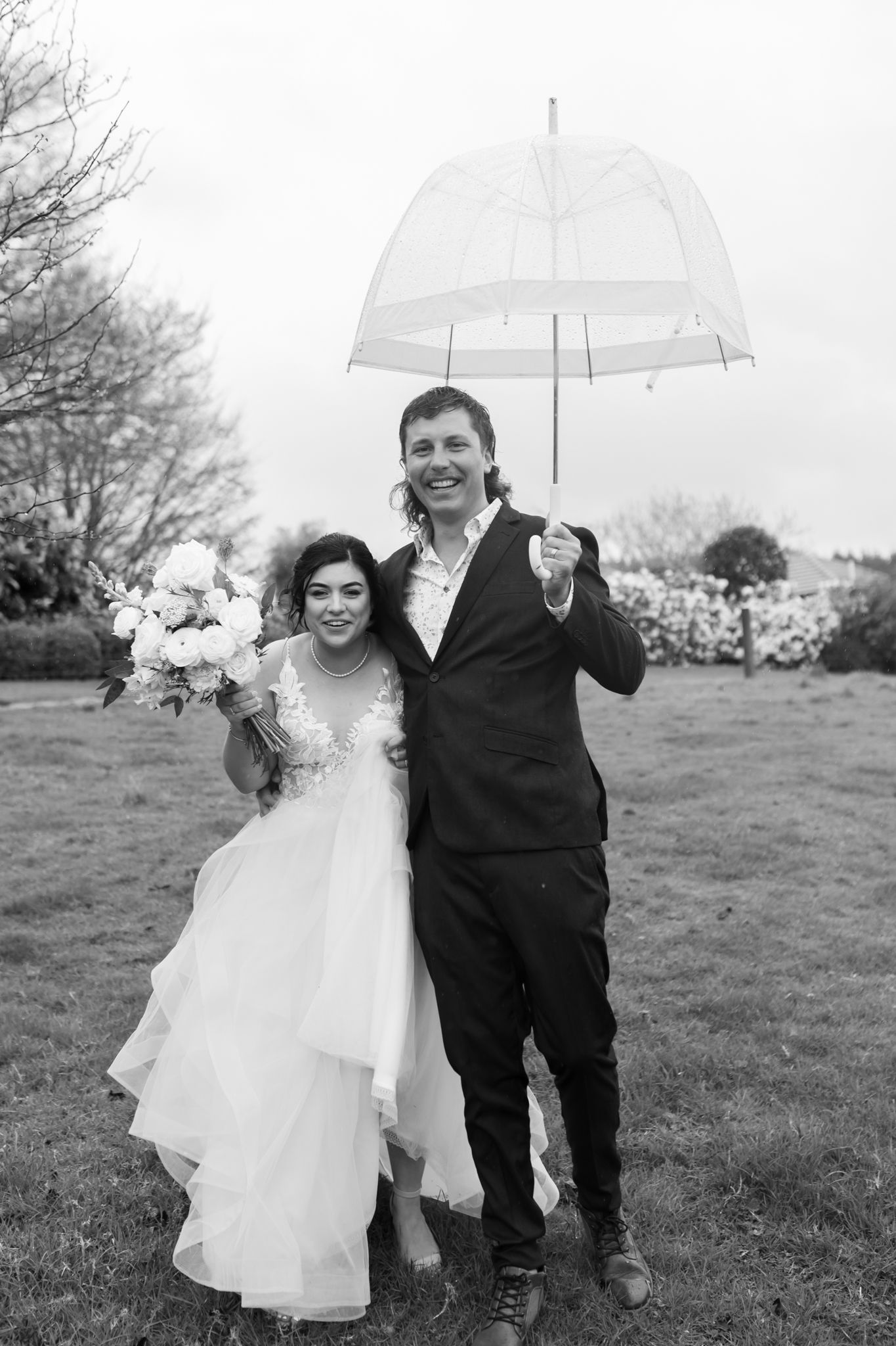 A black and white wedding portrait of a couple sharing an umbrella on a grassy hillside.