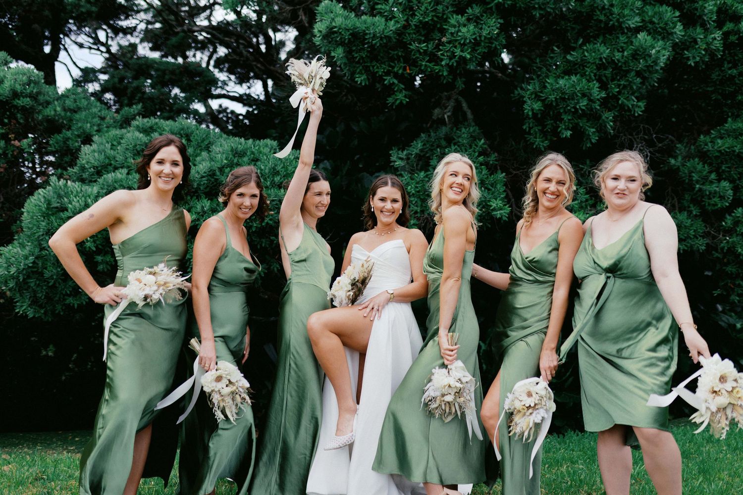Bridesmaids in sage green dresses pose together outdoors for wedding party photos.