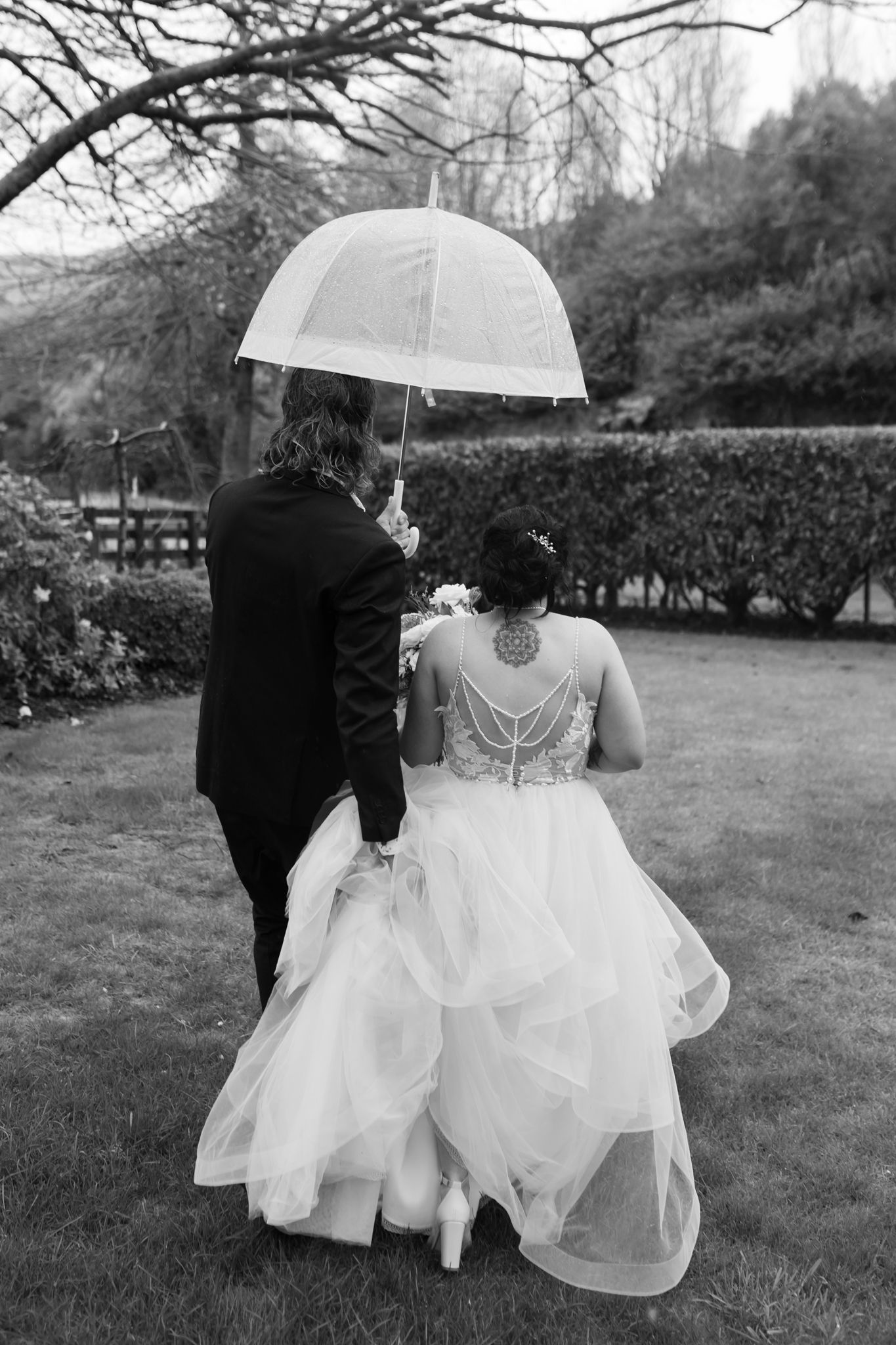 A black and white photo showing the back view of a couple walking with an umbrella on their wedding day.