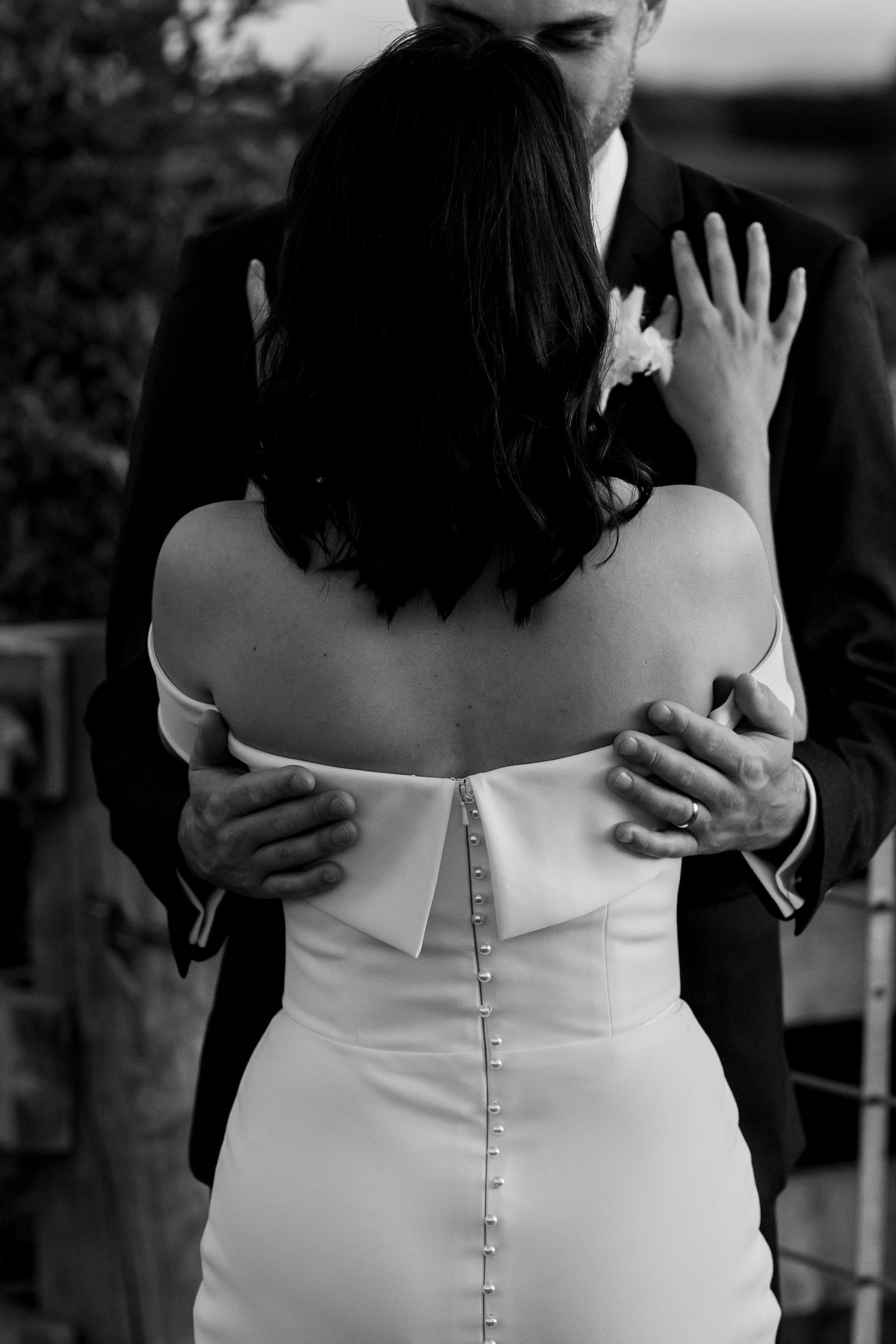 Back view of a bride in a fitted white dress with button details embracing her partner.