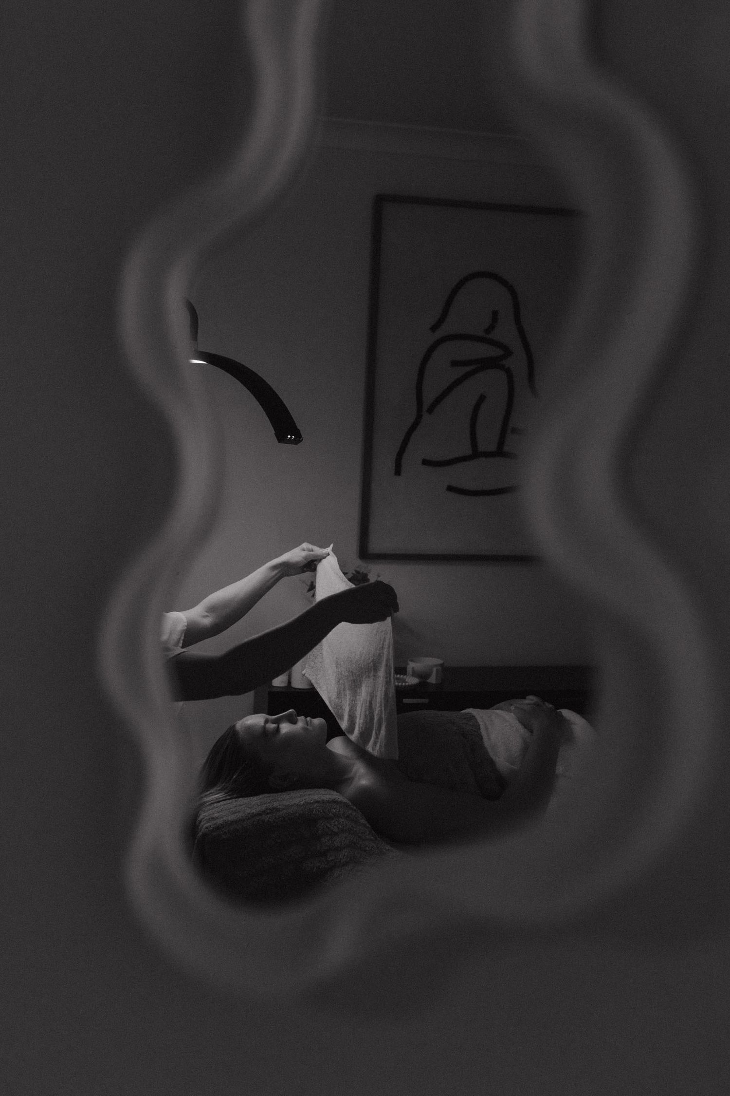Black and white artistic photograph of a person reclining in bed with a hazy, dreamlike effect and wall art in background.