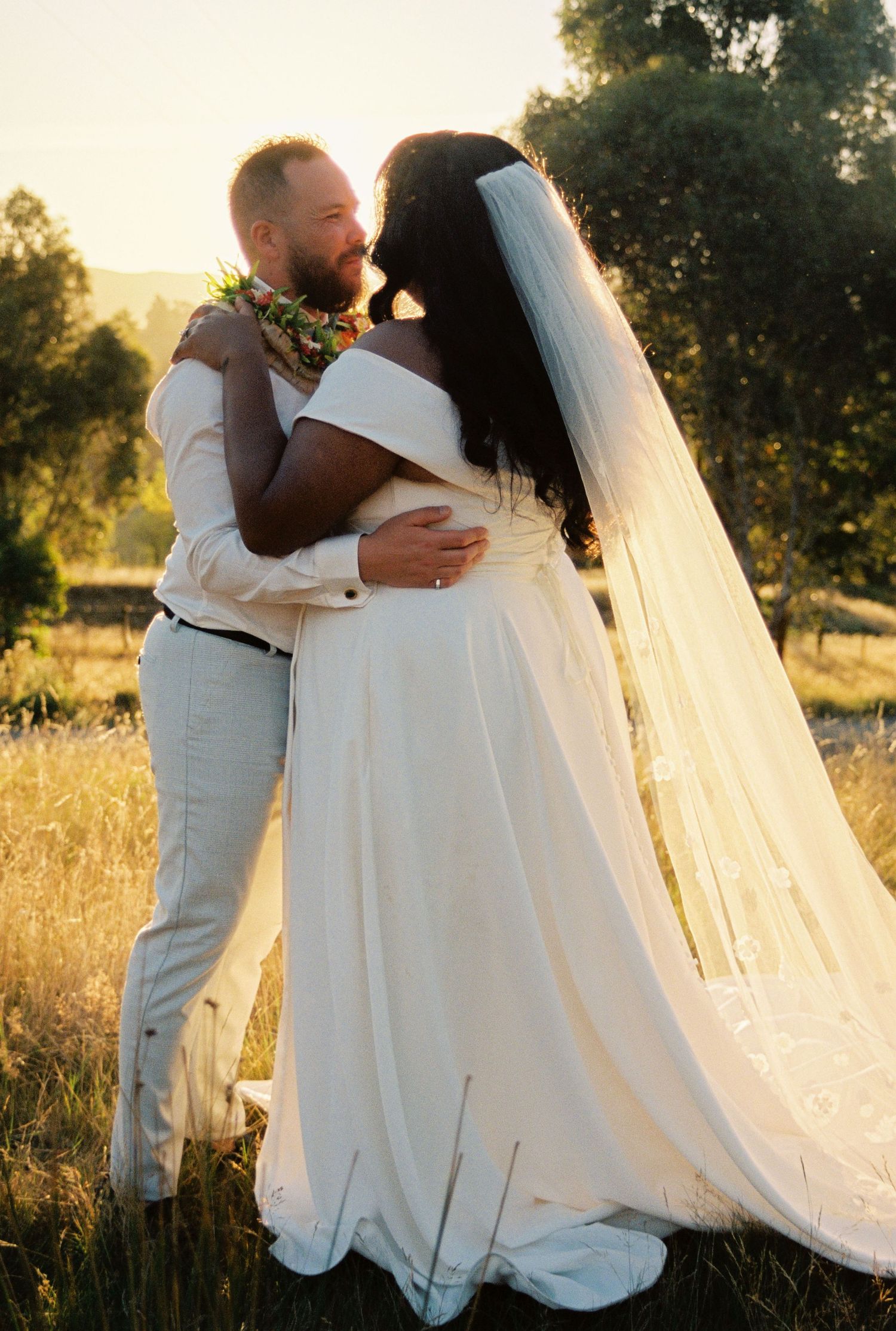 A romantic sunset wedding moment featuring a couple embracing outdoors in a field with golden light and a flowing veil.