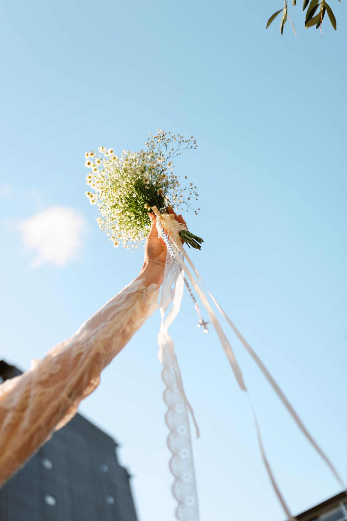 A bride raises a white daisy bouquet with ribbon streamers against a clear blue sky.