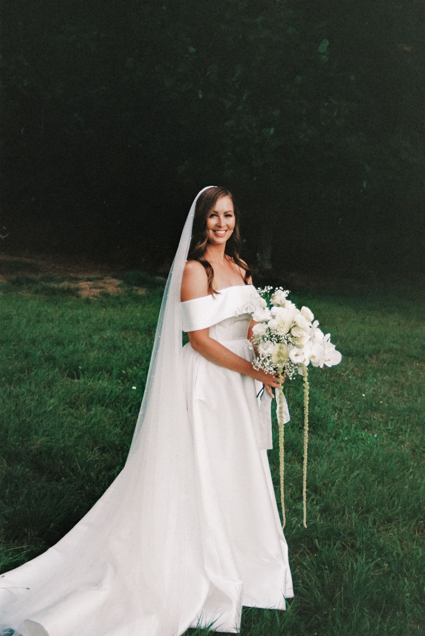 A bride in an off-shoulder white wedding gown holds a lush bouquet of white flowers on a grassy lawn.