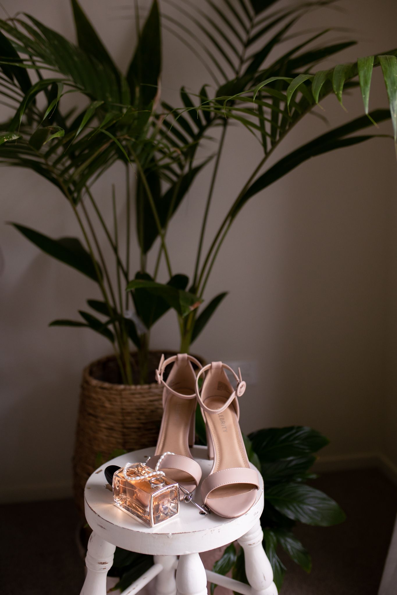 Elegant nude heels and perfume bottle arranged on white side table with tropical plant backdrop.