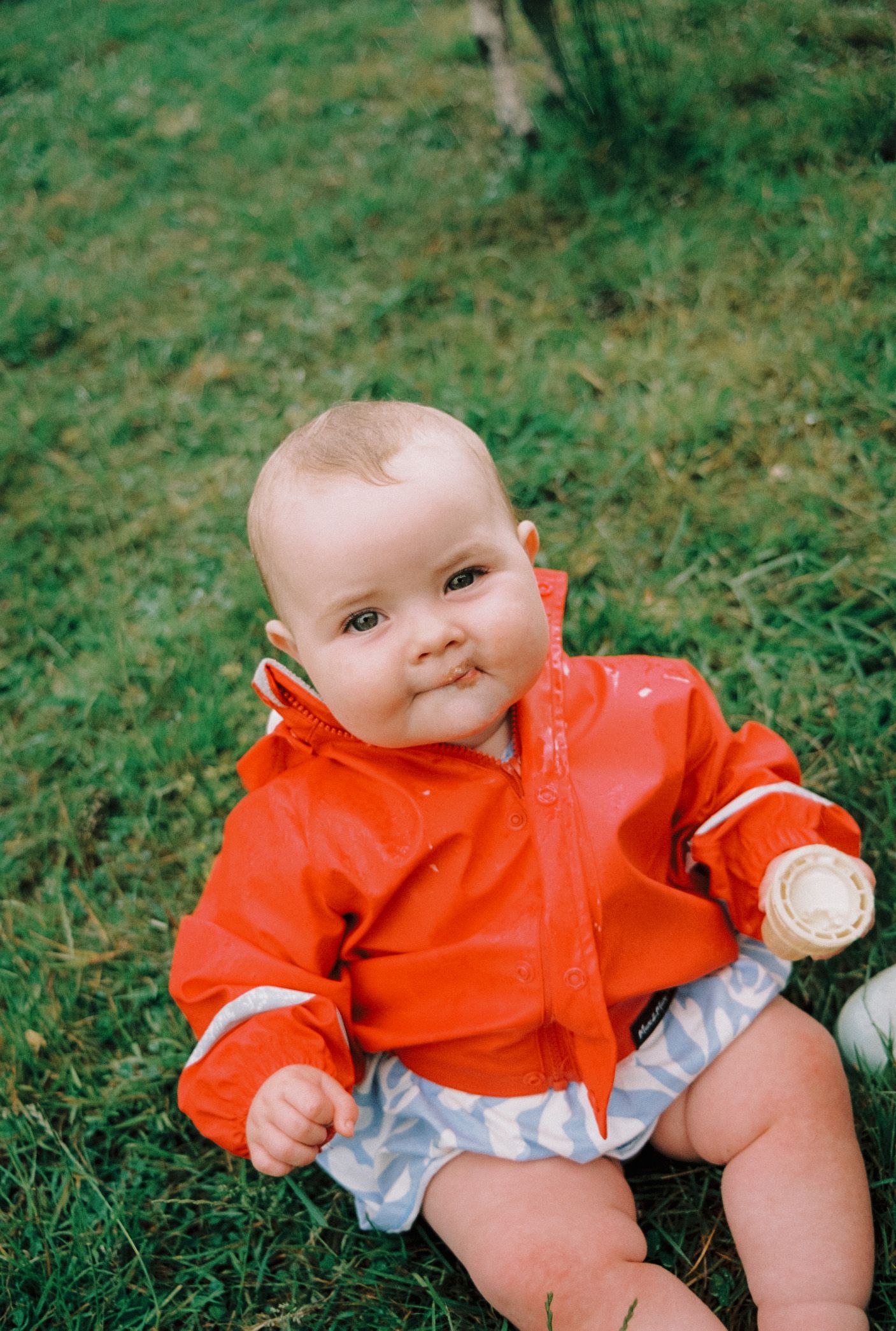 A baby wearing a bright orange raincoat lies on green grass in a sequence of playful poses.
