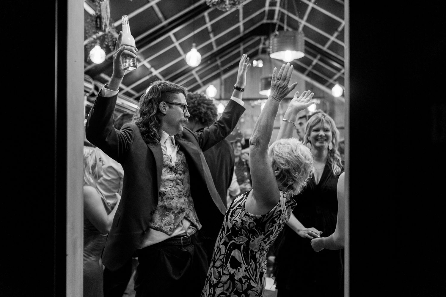 A black and white photo of wedding guests dancing under string lights at an evening reception.