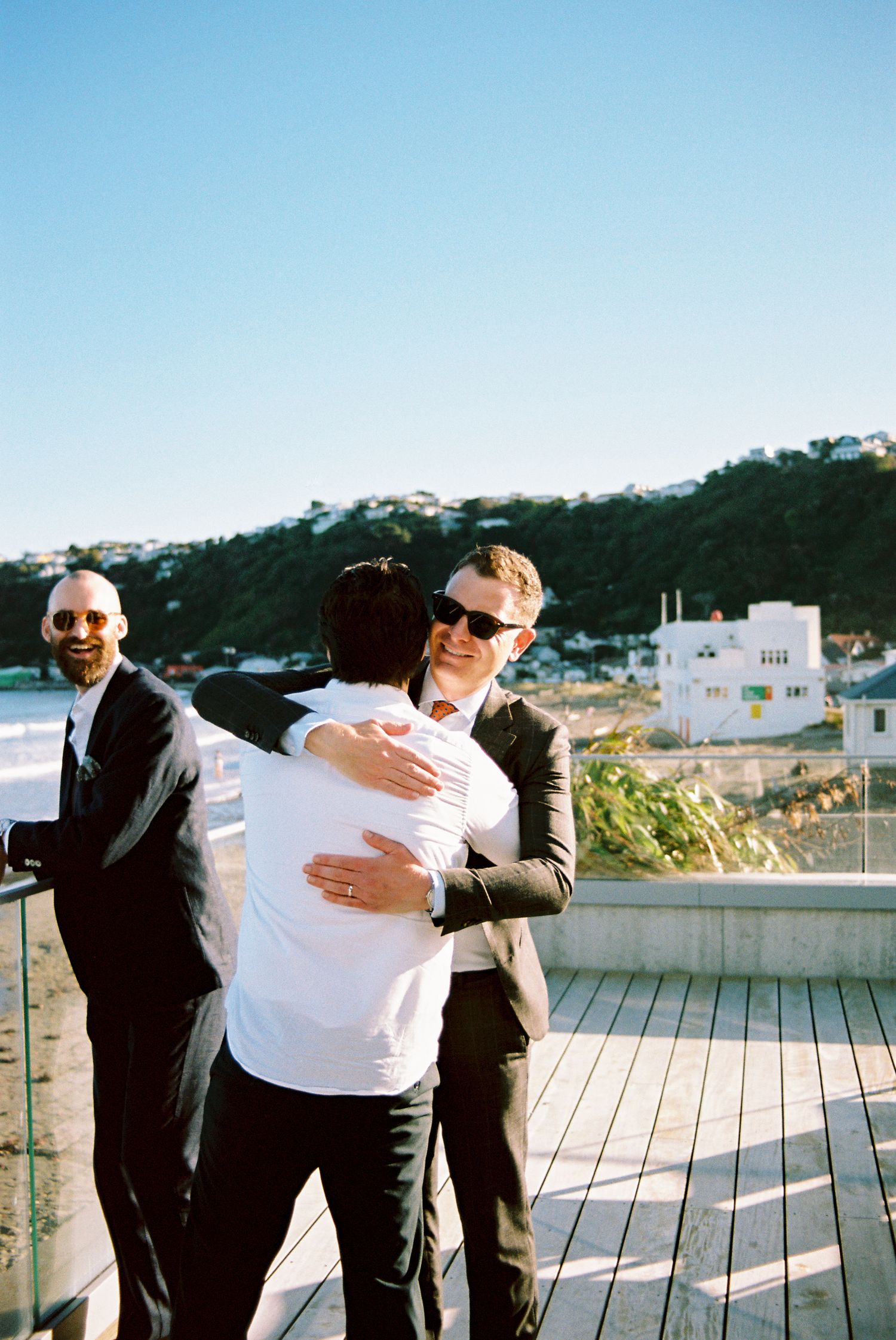 Two people in formal attire share an emotional embrace on an outdoor deck with coastal buildings in the background.
