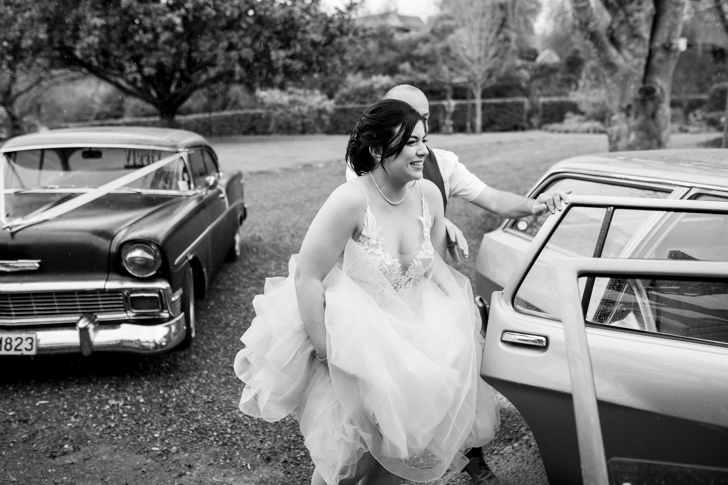 A bride in a flowing white dress getting into a vintage car on her wedding day.