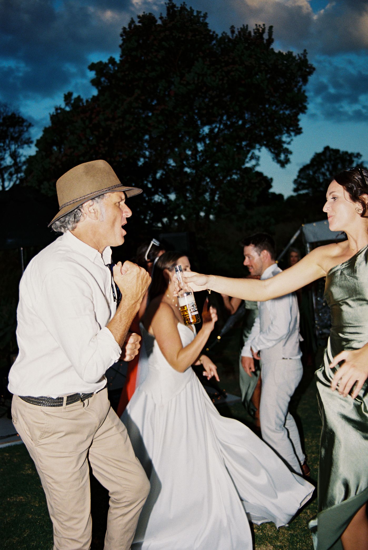 People dancing and celebrating at an outdoor wedding reception under a dusky sky with trees in the background.