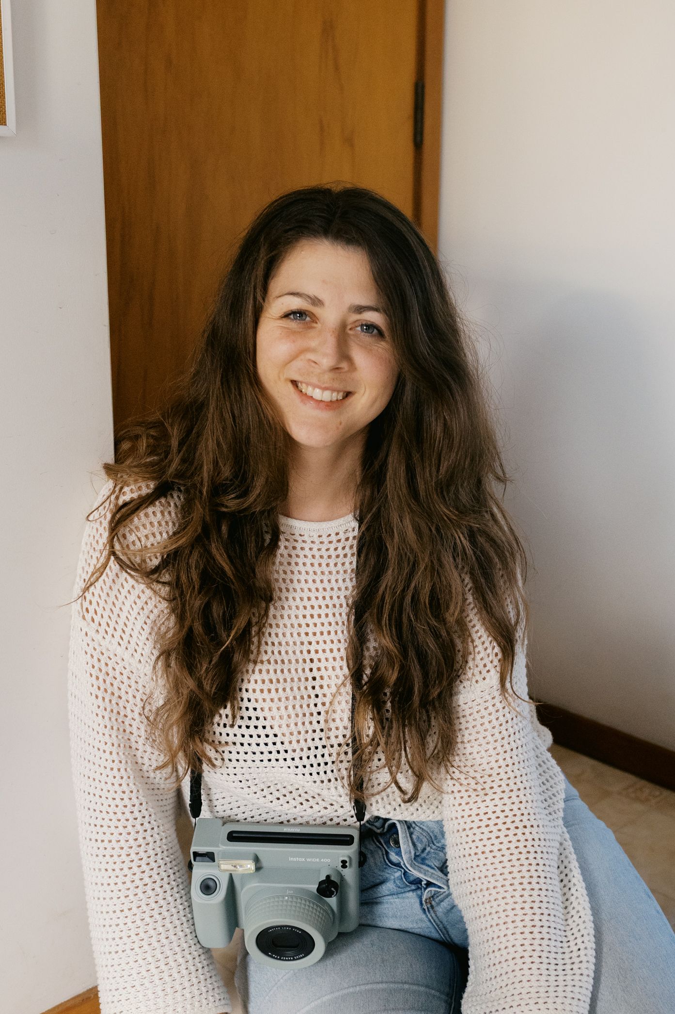 A person in a white knit sweater and denim sits casually indoors with long wavy dark hair.