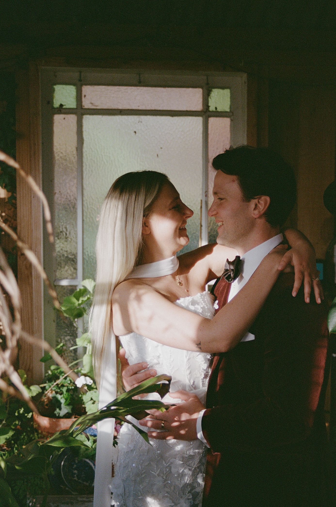 A romantic wedding couple embraces near a window in a dimly lit room with natural light streaming in.