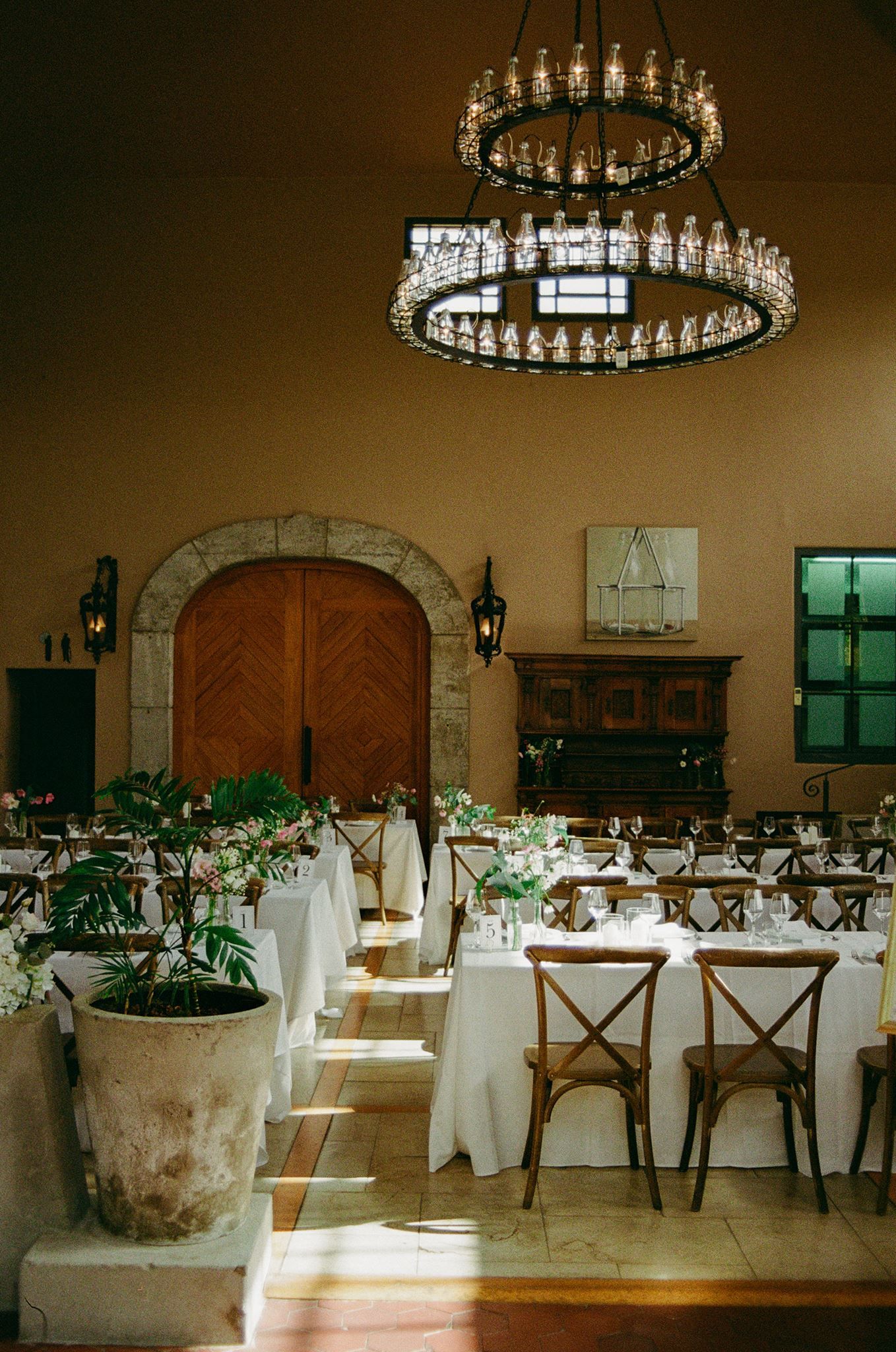 Elegant wedding reception hall with crystal chandeliers, white linen tables, wooden cross-back chairs, and a rustic stone archway.