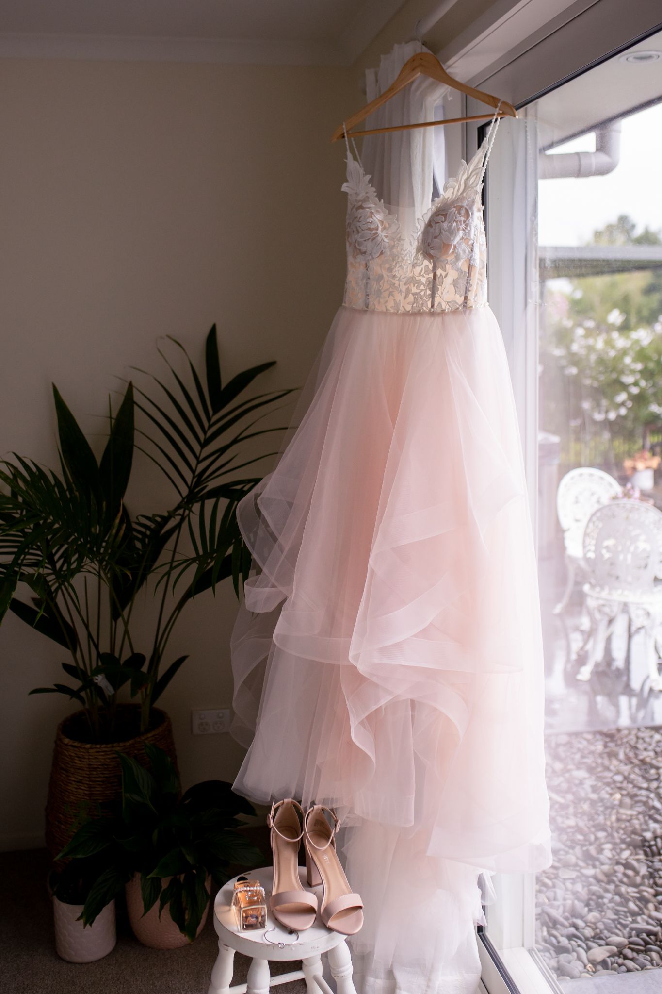 A blush pink wedding gown with lace bodice hanging by a window with bridal shoes below.