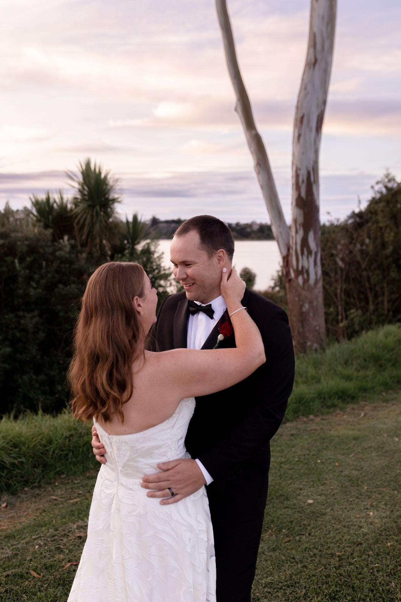 A couple embraces during sunset at their outdoor wedding with trees and water in the background.