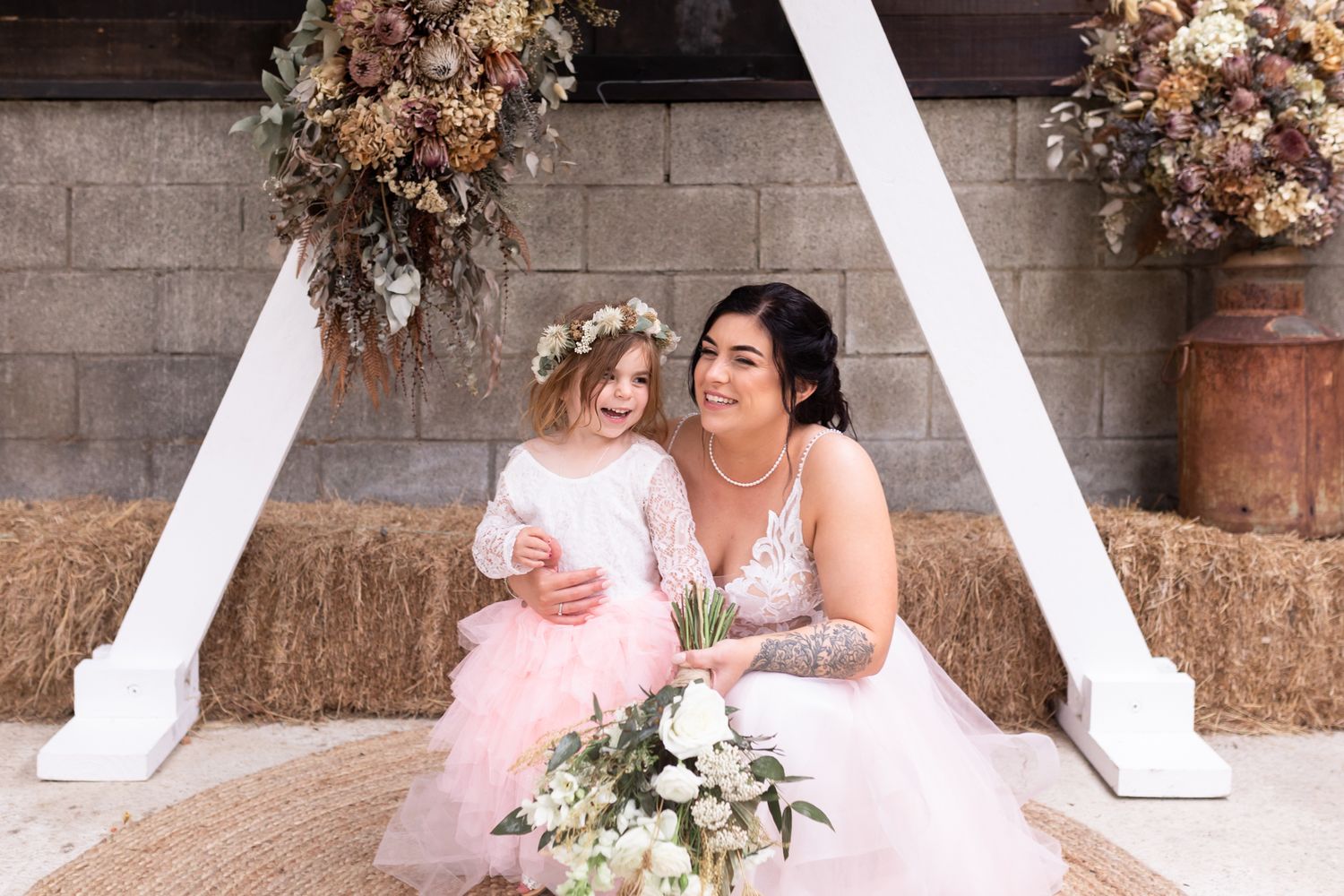 Two brides in pink gowns sit together under floral decorations and white archway.