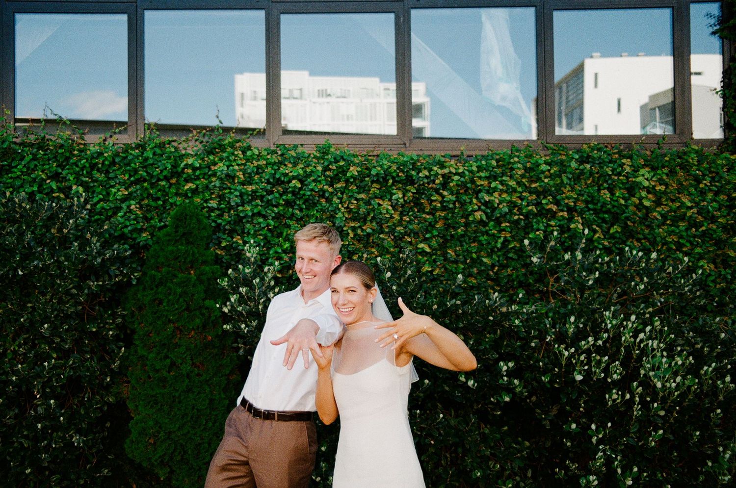 Happy bride and groom posing playfully in front of a lush green ivy wall at their outdoor wedding venue.