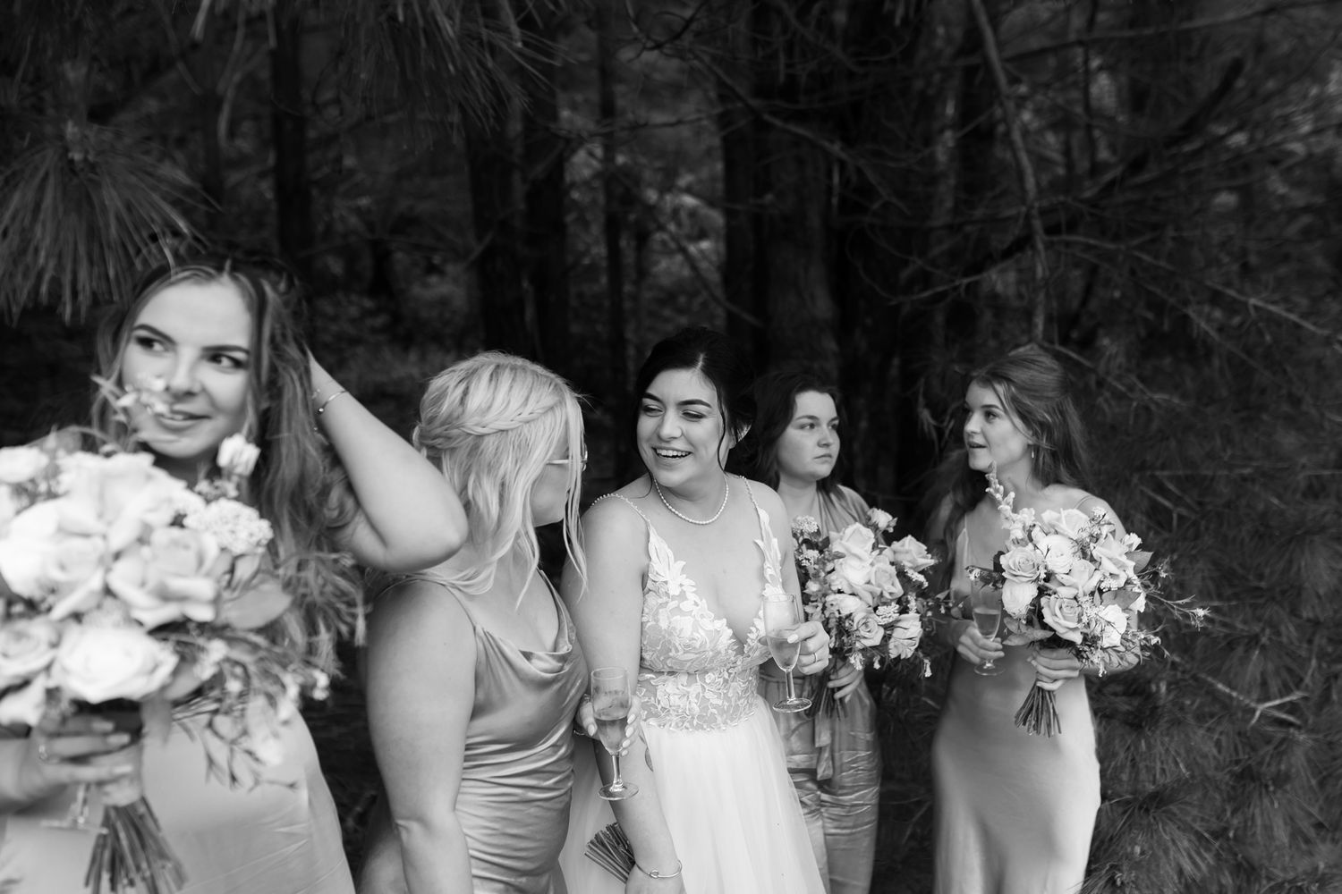 Black and white candid moment of bridal party with bouquets against forest backdrop.