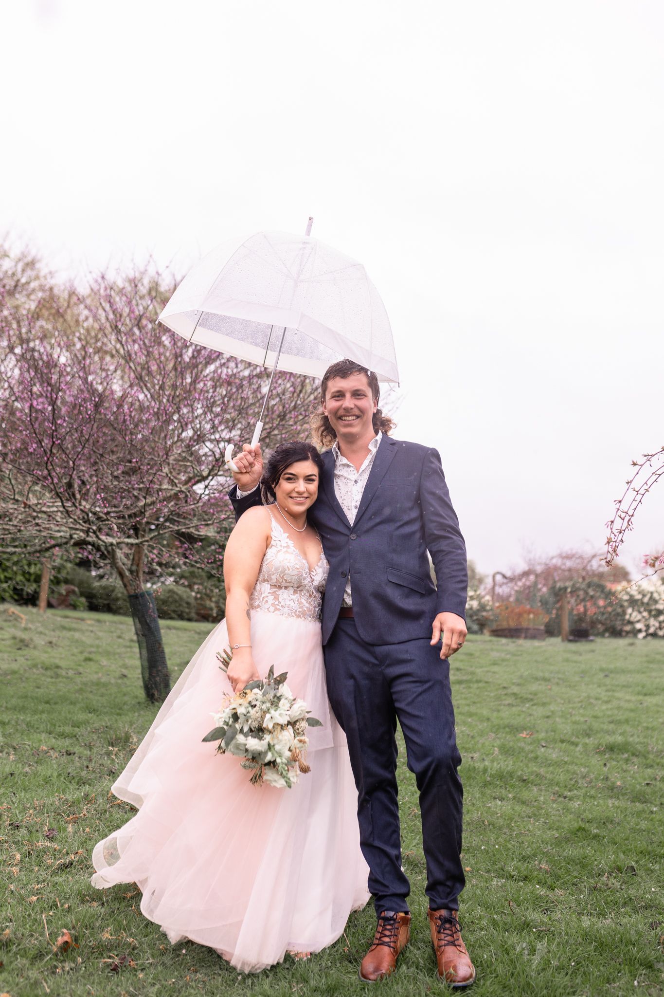 A wedding couple poses outdoors with a clear umbrella against blooming cherry trees in a natural setting.