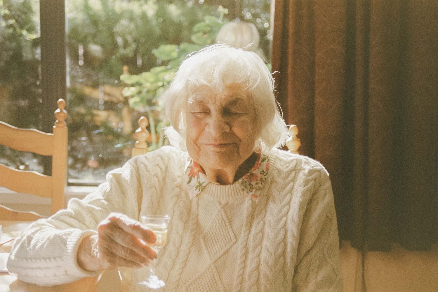 An elderly person in a white sweater smiling while seated at a table.