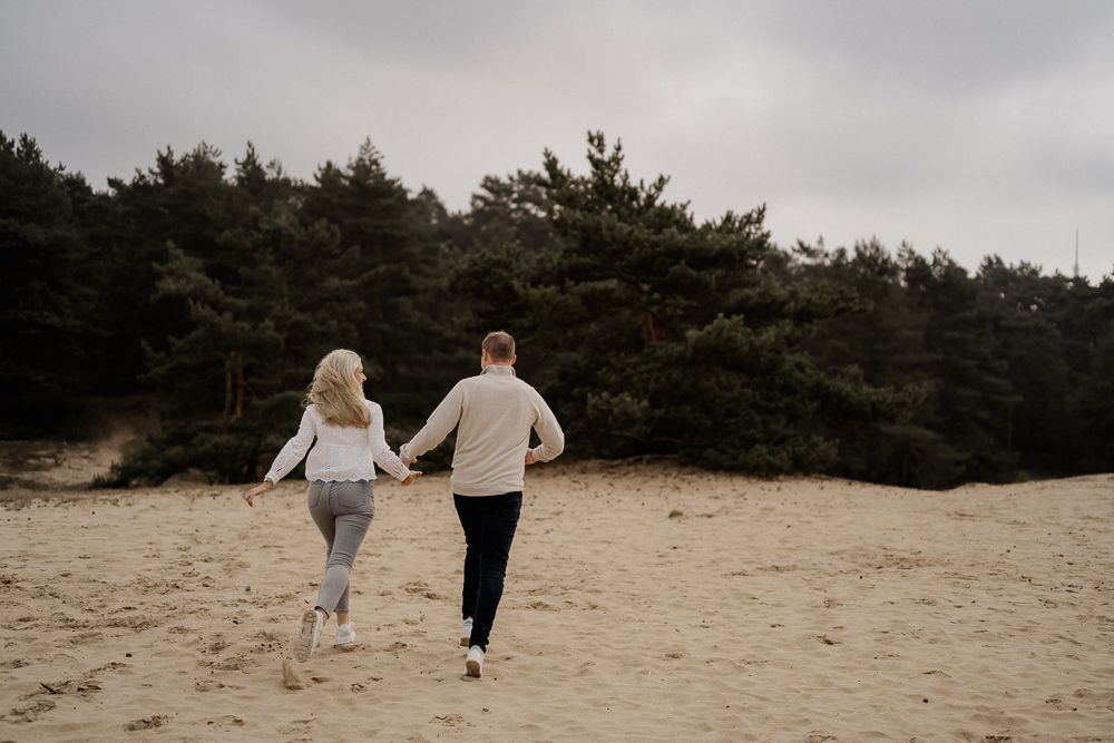 Two people hold hands while running together on a sandy beach with pine forest in the background on a cloudy day. Paarfotos Fotoshooting mit