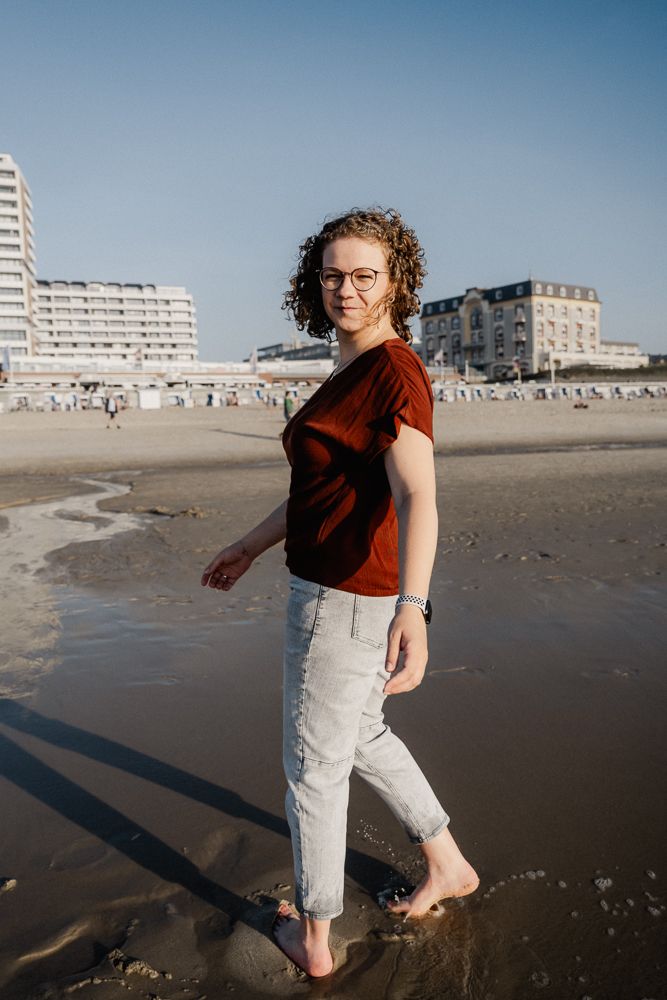 A person in glasses wearing a red t-shirt and light jeans walks barefoot on a beach with buildings in the background.