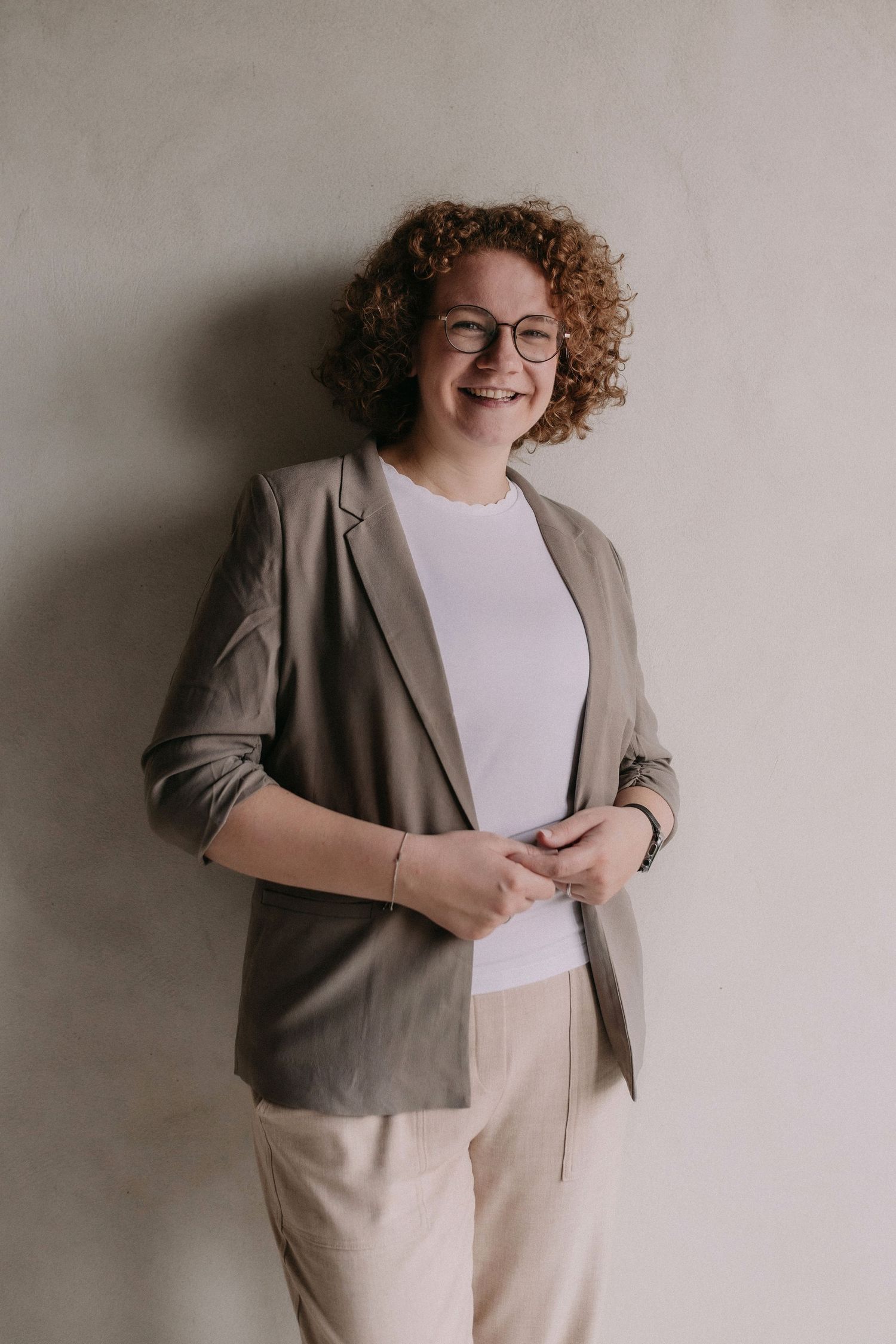 Professional wearing glasses, beige blazer and white top smiles warmly against neutral background.