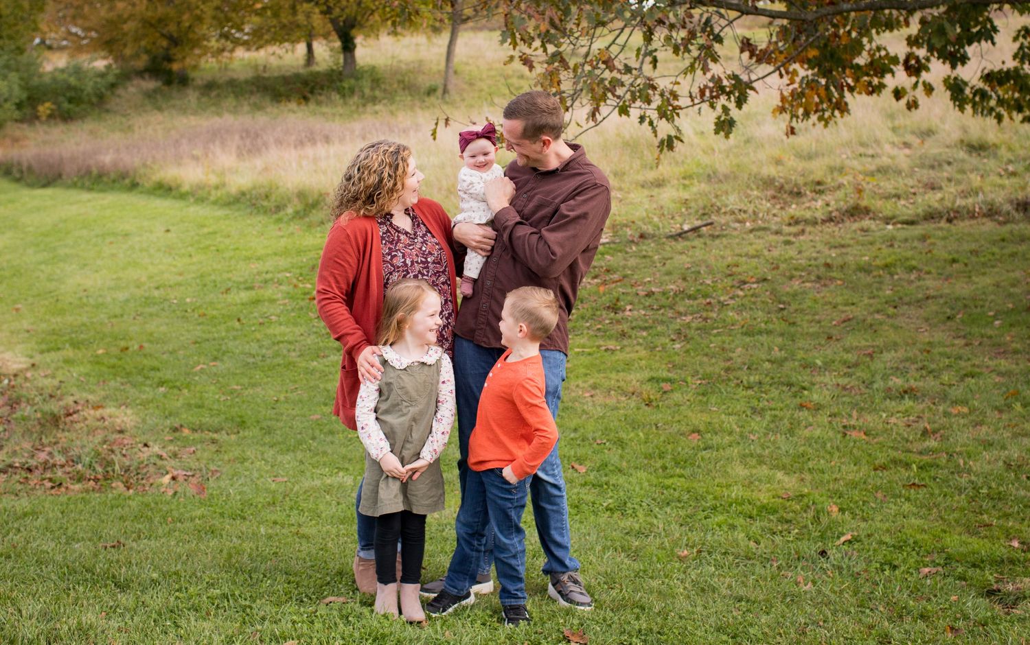 Family of five posing together in a grassy autumn field with colorful trees.