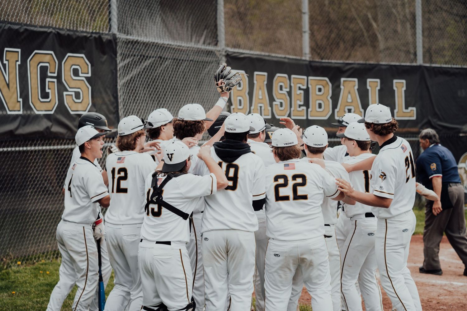 Baseball team in white jerseys huddles together near dugout fence during game.