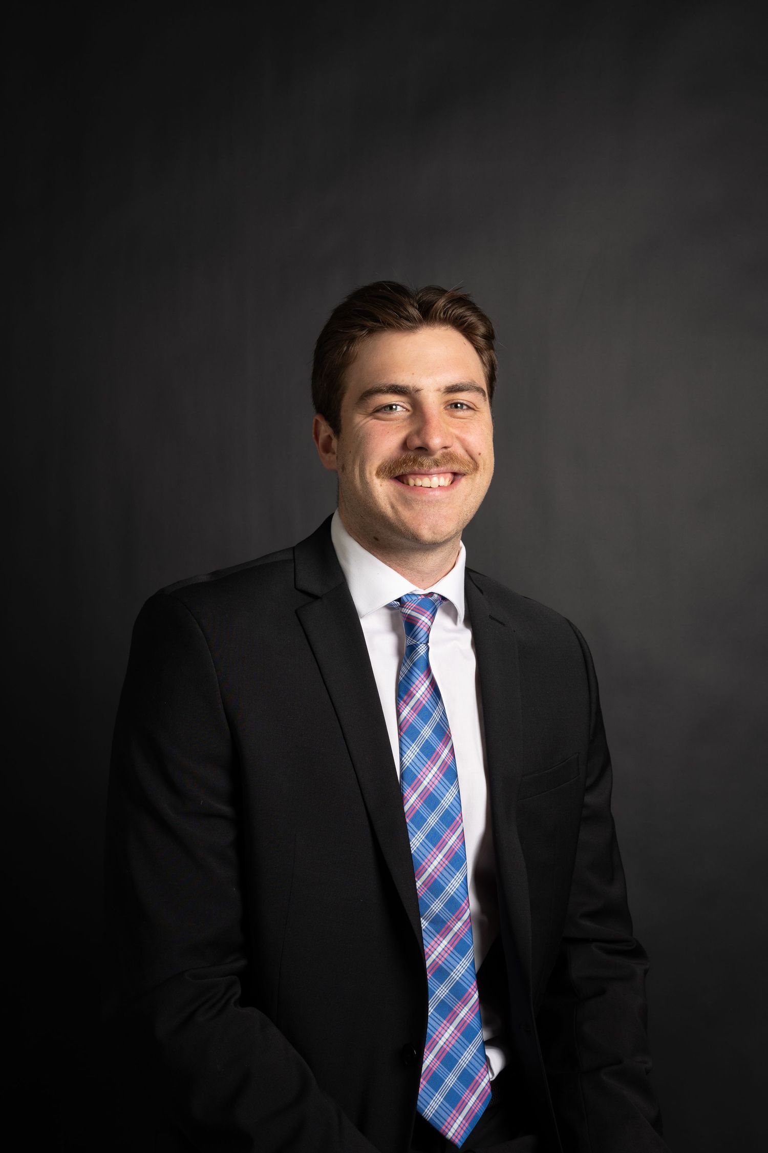 Professional headshot portrait of someone in a black suit with a blue and pink plaid tie against a dark background.