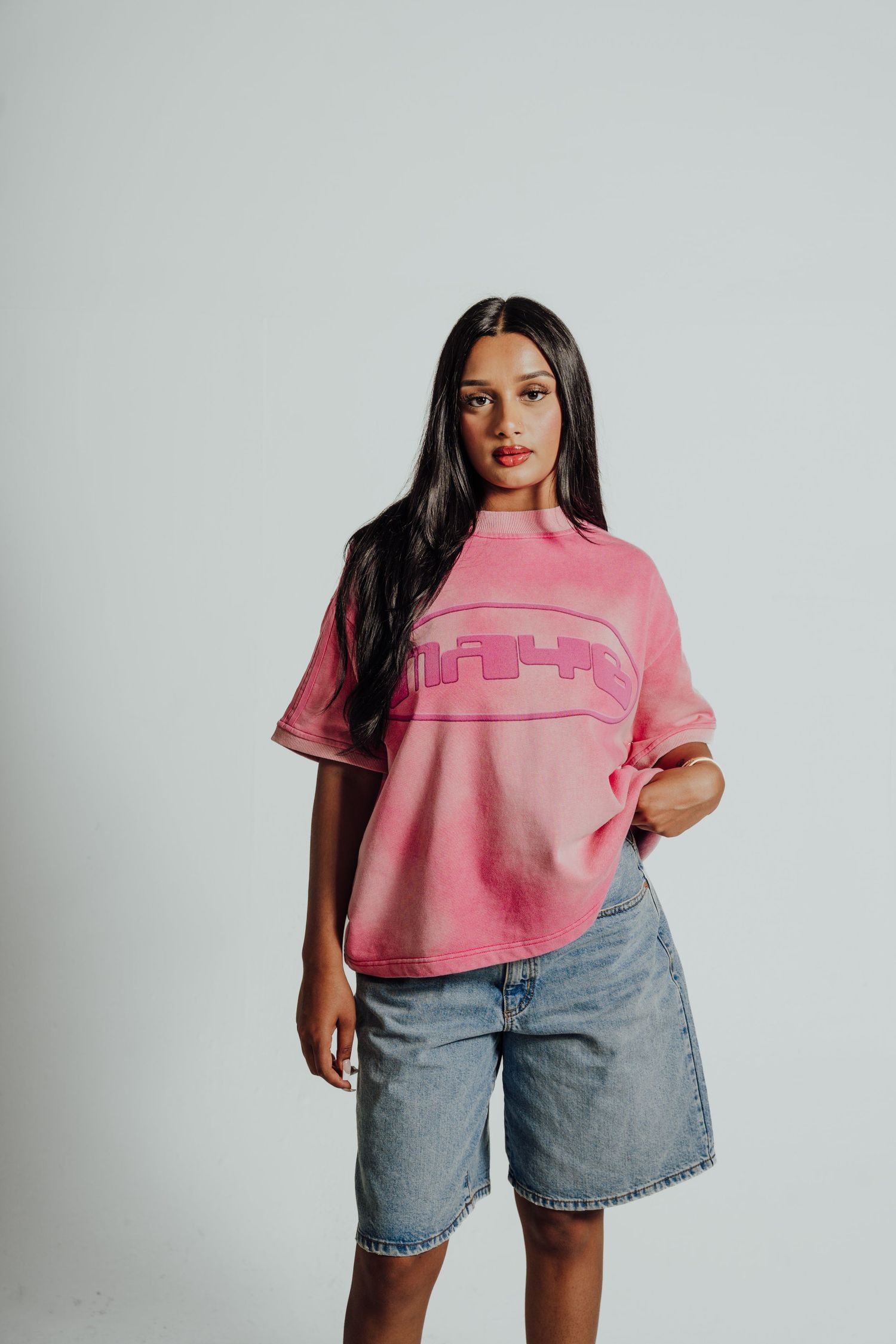 Model wearing a pink cropped t-shirt and light wash denim shorts poses against white background.