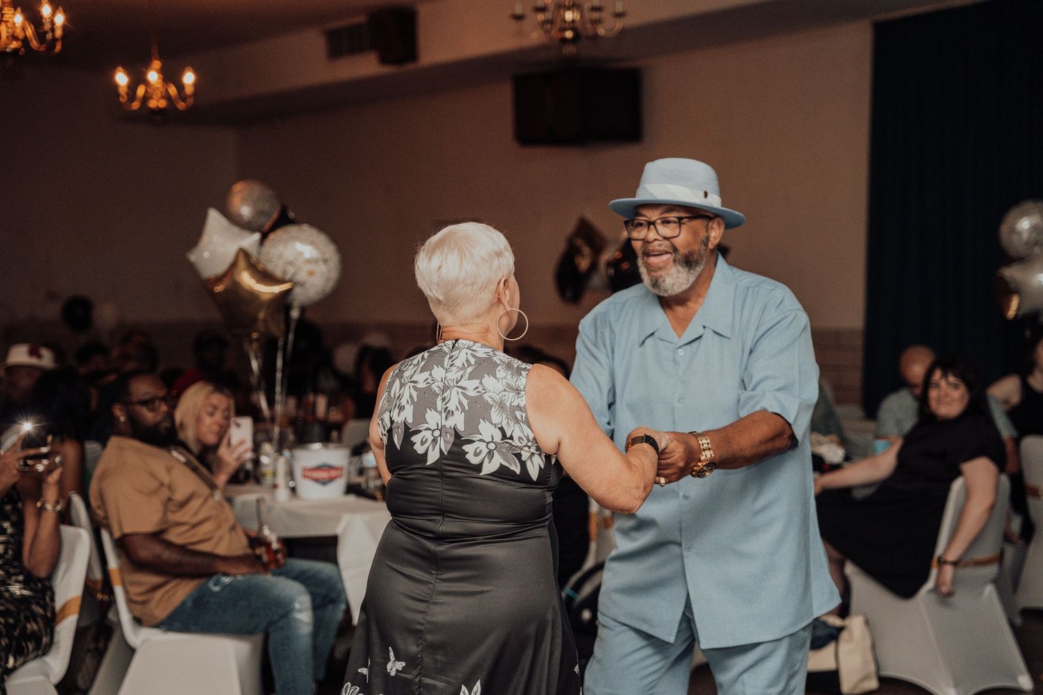 People dancing and celebrating at an indoor event with balloons and chandeliers.