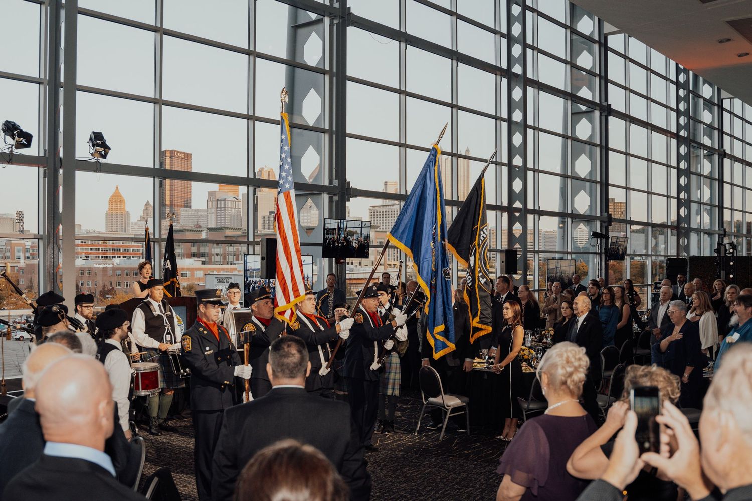 A formal gathering with flags and attendees in a modern venue with floor-to-ceiling windows overlooking a city skyline.