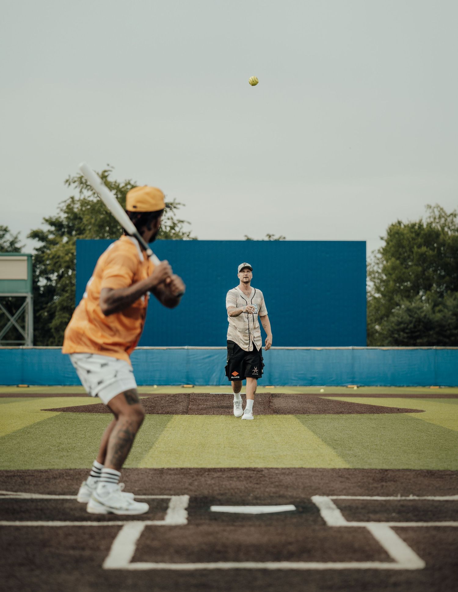 Players on a baseball field practice pitching and catching during a sunny day with a bright blue wall in the background.