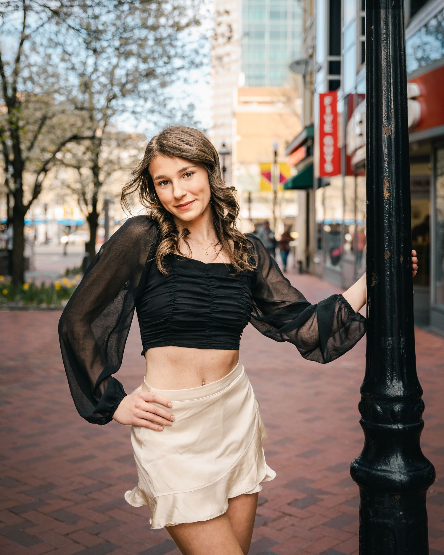 Stylish figure in black sheer-sleeved crop top and flowy beige skirt poses on urban brick street near vintage lamppost.